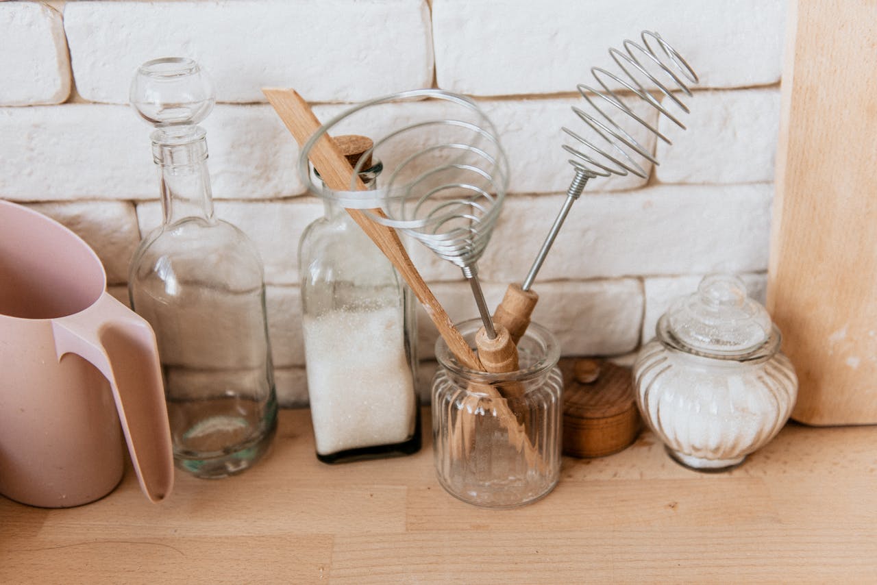our-experience A collection of glass jars and kitchen utensils on a rustic wooden counter with a white brick background.