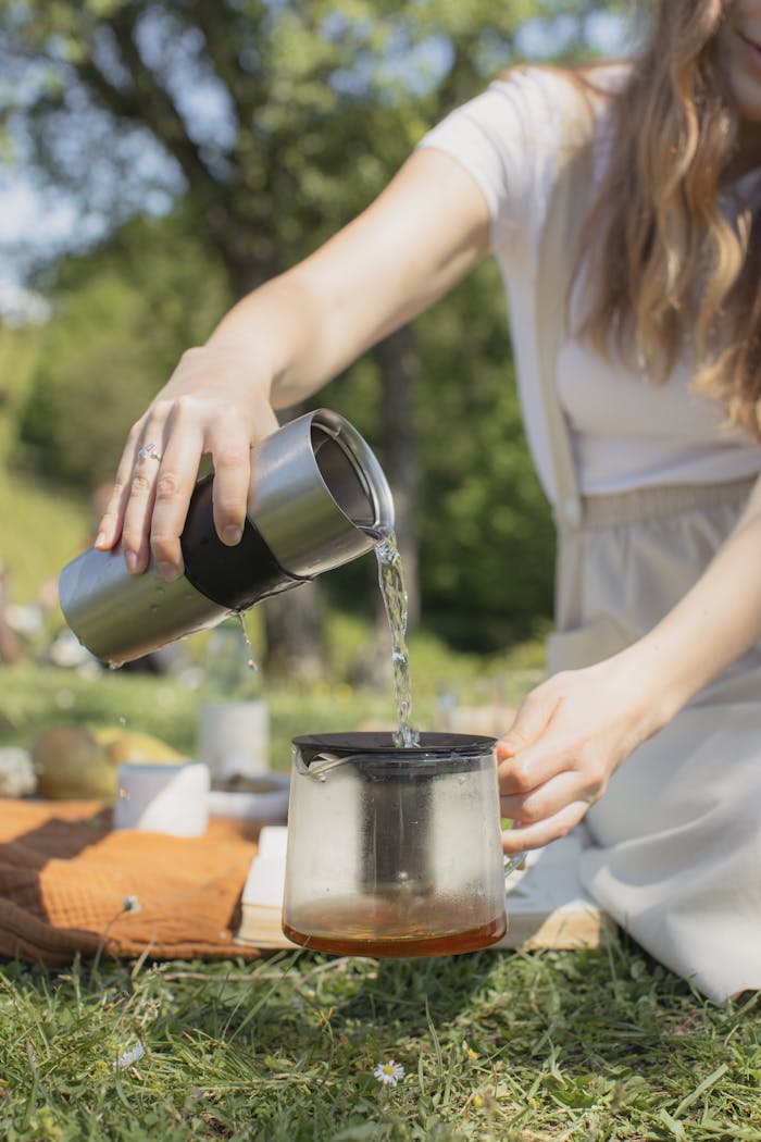 Woman pours tea from a stainless steel tumbler into a glass teapot amidst a grassy outdoor picnic.
