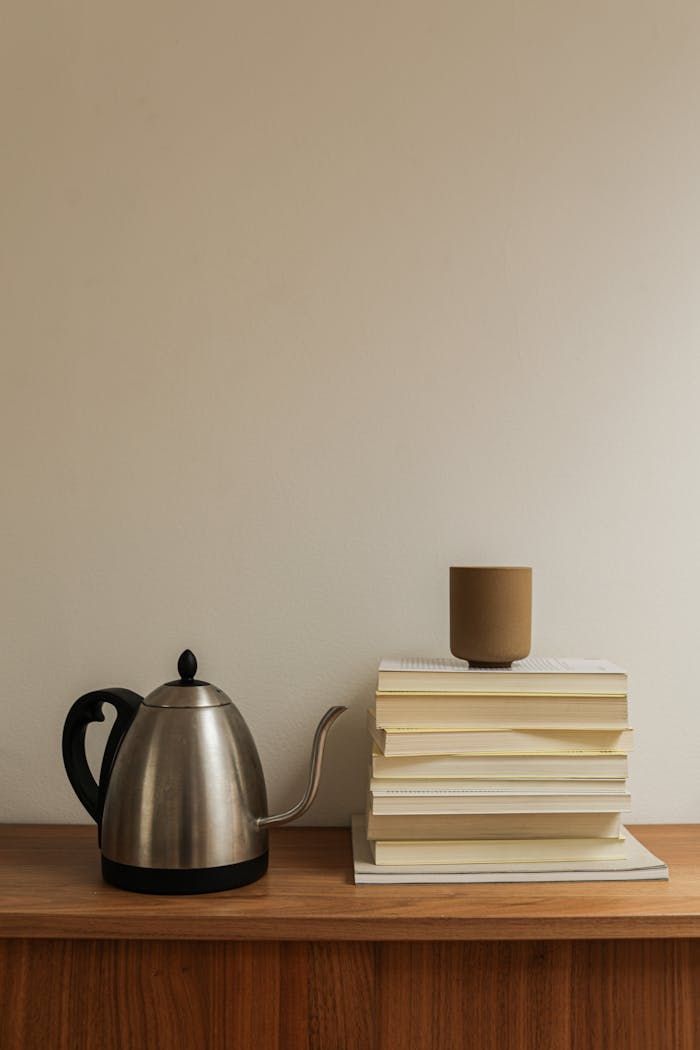 gallery-2 A minimalist arrangement featuring a kettle, ceramic cup, and stacked books on a wooden surface.