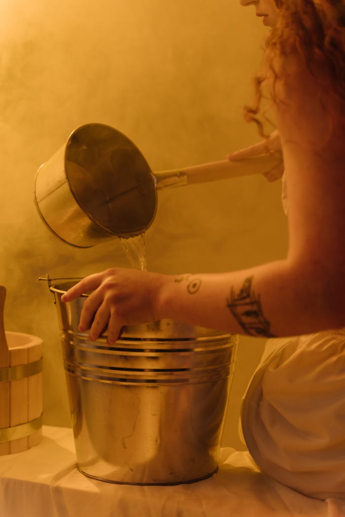 A woman with a tattoo pours water into a stainless steel bucket in a steamy setting.